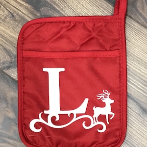 May include: Red quilted oven mitt with a white letter L and a white reindeer design. The reindeer is pulling a sleigh with a scroll design.