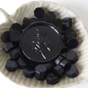 May include: A collection of black wax seal beads and a large, round wax seal with a floral design, presented in a leaf-shaped ceramic dish. The seals are a deep, glossy black.