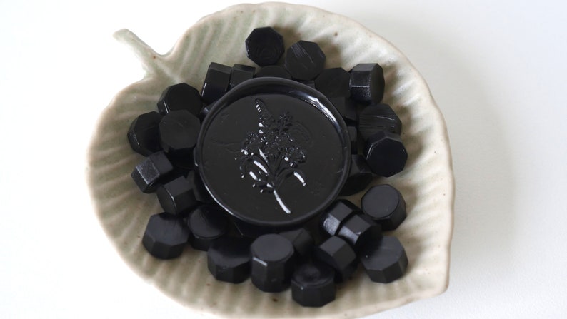 May include: A collection of black wax seal beads and a large, round wax seal with a floral design, presented in a leaf-shaped ceramic dish. The seals are a deep, glossy black.