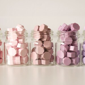 May include: Five small glass jars filled with colorful wax seals in shades of pink and purple. The seals have a geometric shape and are arranged in a gradient of colors, from light pink to lavender. The jars are clear, and the background is white.