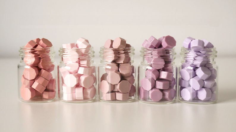 May include: Five small glass jars filled with colorful wax seals in shades of pink and purple. The seals have a geometric shape and are arranged in a gradient of colors, from light pink to lavender. The jars are clear, and the background is white.