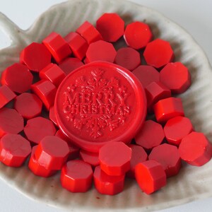 May include: A collection of red wax seals and sealing wax pieces in a leaf-shaped dish. The central seal is embossed with "MERRY CHRISTMAS" and a snowflake design. The wax pieces are various geometric shapes.