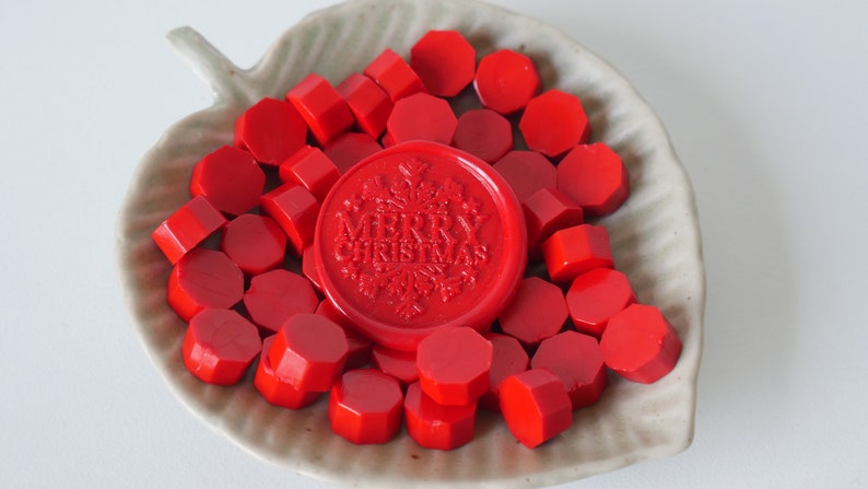 May include: A collection of red wax seals and sealing wax pieces in a leaf-shaped dish. The central seal is embossed with "MERRY CHRISTMAS" and a snowflake design. The wax pieces are various geometric shapes.