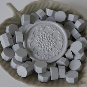 May include: A collection of gray wax seal beads and a decorative seal stamp with a floral design. The items are arranged in a leaf-shaped ceramic dish. The seal stamp features a detailed floral pattern, and the beads are hexagonal.