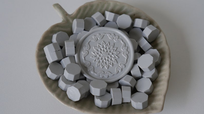 May include: A collection of gray wax seal beads and a decorative seal stamp with a floral design. The items are arranged in a leaf-shaped ceramic dish. The seal stamp features a detailed floral pattern, and the beads are hexagonal.