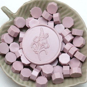 May include: A collection of light purple wax seal beads and a round seal with a floral design, presented in a leaf-shaped ceramic dish. The beads are hexagonal. The seal has a floral design.