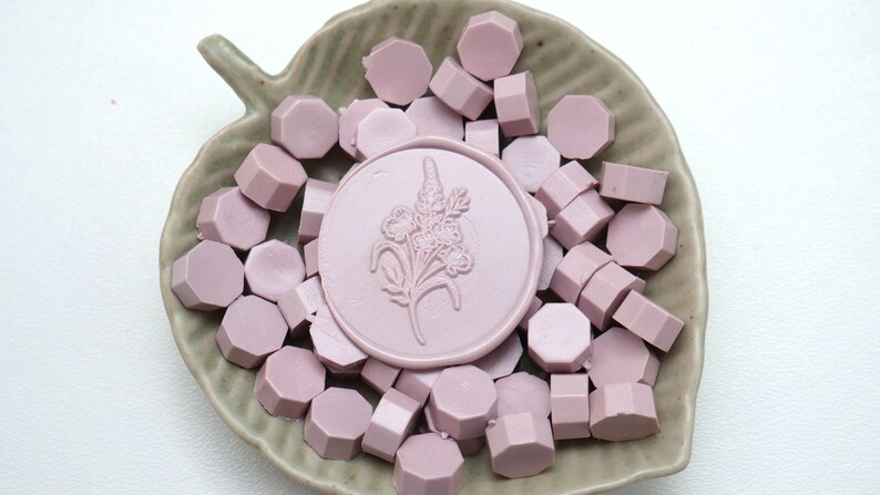 May include: A collection of light purple wax seal beads and a round seal with a floral design, presented in a leaf-shaped ceramic dish. The beads are hexagonal. The seal has a floral design.