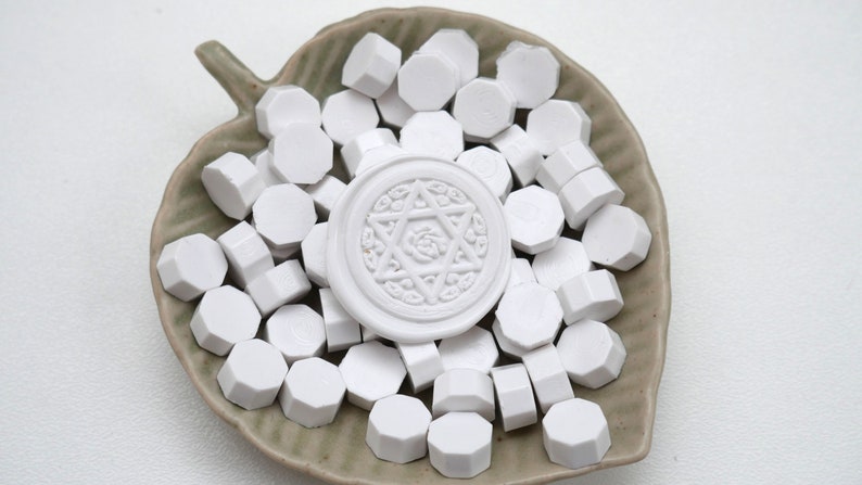Op de afbeelding: Witte lakzegelparels en een witte zegelstempel met een ster- en bloemenontwerp, gepresenteerd op een blad-vormige keramische schaal. De parels zijn zeshoekig.