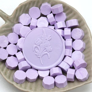 May include: A collection of lavender-colored wax seals and wax seal beads arranged on a leaf-shaped ceramic dish. The central seal features a floral design. The beads are hexagonal in shape.