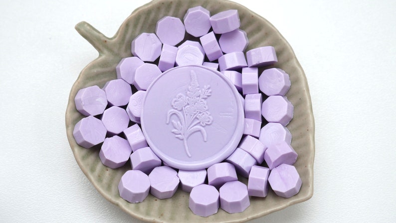 May include: A collection of lavender-colored wax seals and wax seal beads arranged on a leaf-shaped ceramic dish. The central seal features a floral design. The beads are hexagonal in shape.