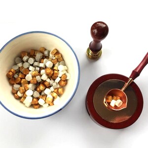 Pode incluir: Kit para fazer selos de cera com contas de cera douradas, brancas e cinzentas, um selo com cabo de madeira e uma colher para derreter cera. Os artigos estão dispostos sobre uma superfície branca.