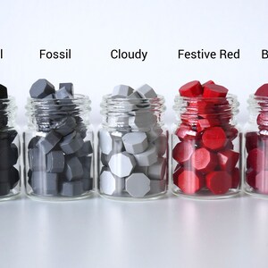 May include: Five small glass jars filled with colorful wax seals. The colors are Charcoal, Fossil, Cloudy, Festive Red, and Burgundy. Each jar is labeled with its respective color name in black text. The wax seals have a geometric shape.