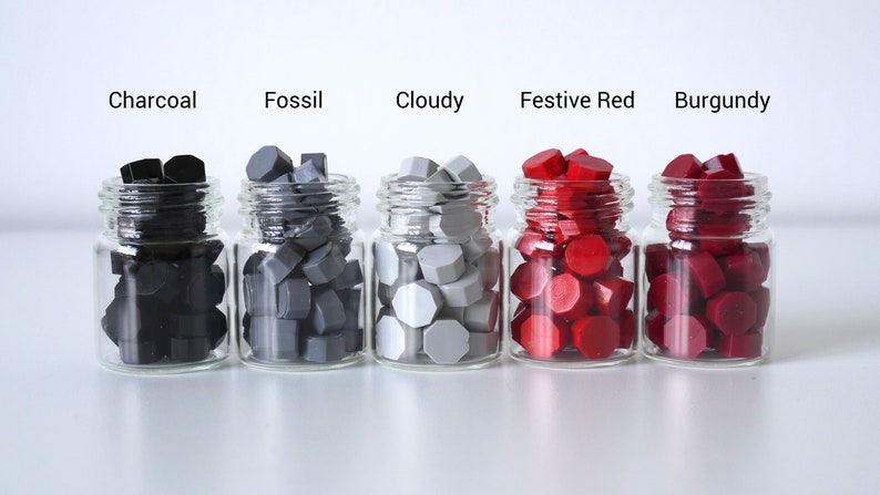 May include: Five small glass jars filled with colorful wax seals. The colors are Charcoal, Fossil, Cloudy, Festive Red, and Burgundy. Each jar is labeled with its respective color name in black text. The wax seals have a geometric shape.