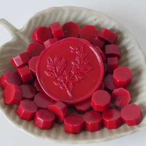 May include: A collection of red sealing wax pieces and a round wax seal with a floral design, presented in a leaf-shaped ceramic dish. The wax pieces are hexagonal and the seal is embossed with a detailed botanical image.