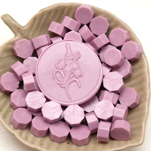 May include: A collection of lavender-colored wax seal beads and a large round seal with a floral design. The seals are arranged in a leaf-shaped ceramic dish. The wax beads are hexagonal in shape.