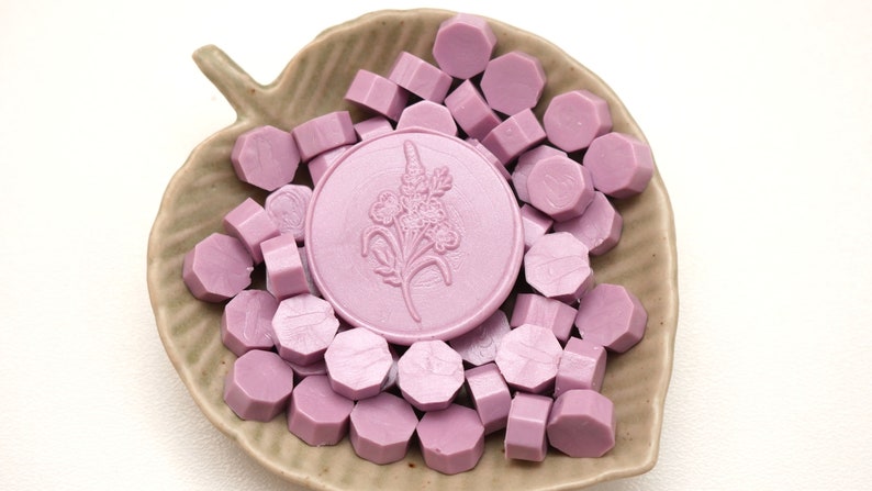 May include: A collection of lavender-colored wax seal beads and a large round seal with a floral design. The seals are arranged in a leaf-shaped ceramic dish. The wax beads are hexagonal in shape.