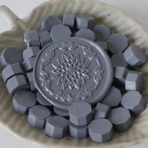 May include: A collection of gray wax seal beads and a large seal, arranged in a leaf-shaped ceramic dish. The seal features a detailed floral design. The beads are hexagonal in shape.