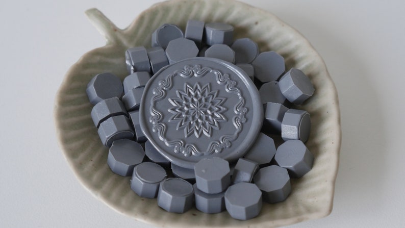 May include: A collection of gray wax seal beads and a large seal, arranged in a leaf-shaped ceramic dish. The seal features a detailed floral design. The beads are hexagonal in shape.