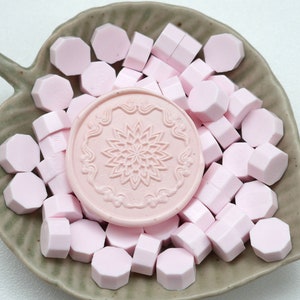May include: A collection of pink wax seal beads and a pink wax seal stamp with a floral design, presented on a leaf-shaped ceramic dish. The wax beads are hexagonal. The dish is a muted green color.