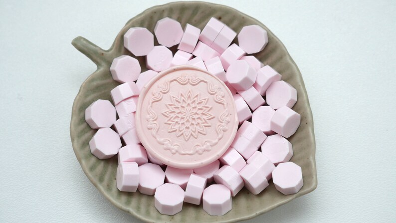 May include: A collection of pink wax seal beads and a pink wax seal stamp with a floral design, presented on a leaf-shaped ceramic dish. The wax beads are hexagonal. The dish is a muted green color.
