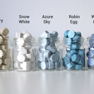 Op de afbeelding: Vijf kleine glazen potjes gevuld met kleurrijke waszegels. De kleuren zijn gelabeld: Ivoor, Sneeuwwit, Azure Sky, Robin Egg en Washed Denim. Elk potje bevat meerdere zeshoekige waszegels.