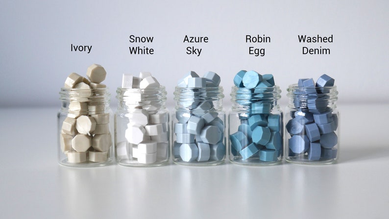Op de afbeelding: Vijf kleine glazen potjes gevuld met kleurrijke waszegels. De kleuren zijn gelabeld: Ivoor, Sneeuwwit, Azure Sky, Robin Egg en Washed Denim. Elk potje bevat meerdere zeshoekige waszegels.