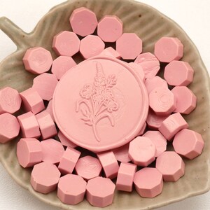 May include: A collection of pink wax seals and wax seal beads arranged in a leaf-shaped dish. The central seal features a floral design. The seals and beads are a soft, muted pink color, creating a delicate and elegant aesthetic.