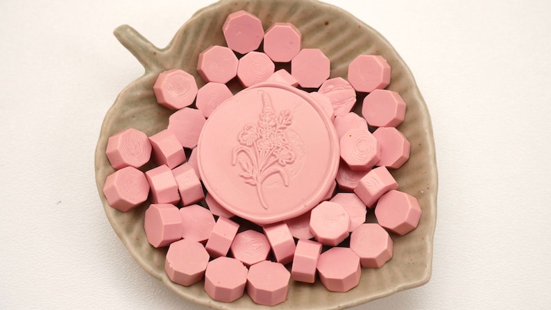 May include: A collection of pink wax seals and wax seal beads arranged in a leaf-shaped dish. The central seal features a floral design. The seals and beads are a soft, muted pink color, creating a delicate and elegant aesthetic.