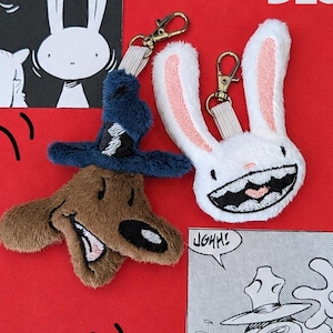 May include: Two plush keychains featuring a brown dog wearing a blue hat and a white rabbit with a wide grin. The keychains include gold-tone metal lobster clasps and are displayed against a red background with black and white comic illustrations.