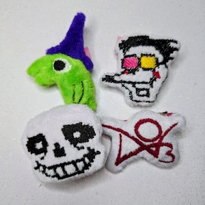 May include: Four plush toys are shown. One is a green creature with a purple hat. Another is a white head with black details. The third is a white head with black details and pink and yellow glasses. The last is a white shape with red details.