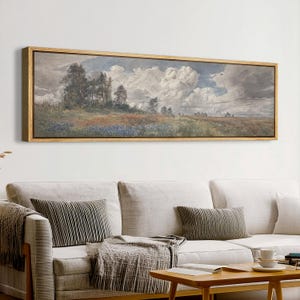 May include: A landscape painting in a rectangular gold frame. The artwork depicts a field of wildflowers with trees under a cloudy sky. The painting is displayed above a white sofa with pillows and a wooden coffee table.
