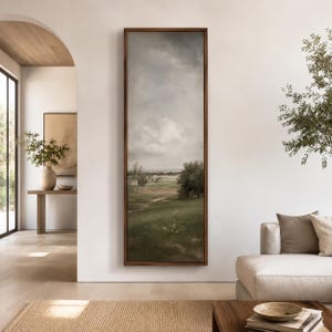 May include: A framed landscape painting with a dark wood frame. The artwork depicts a cloudy sky over a green field with trees. The painting is displayed on a white wall in a well-lit room with neutral tones.