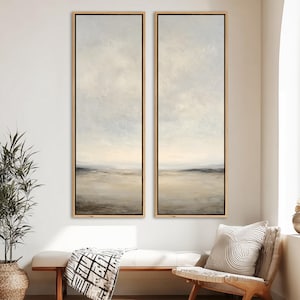 May include: Two vertical framed landscape paintings. The artwork features a muted color palette of beige, gray, and white, with a soft, blurred effect. The frames are a light wood color, and the paintings are displayed in a well-lit room.