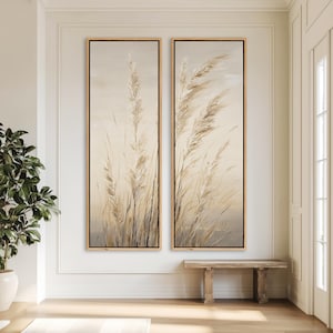 May include: Two vertical framed canvas art pieces. Each features a beige and brown depiction of tall grasses blowing in the wind. The frames are a light wood color with a black border. The artwork is displayed on a white wall.