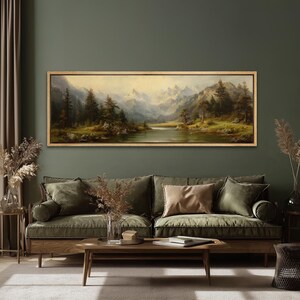 May include: A framed landscape painting depicts a serene mountain scene with a river, trees, and snow-capped peaks. The artwork is encased in a light brown wooden frame and hangs above a green sofa with decorative pillows. The room has a dark green wall.