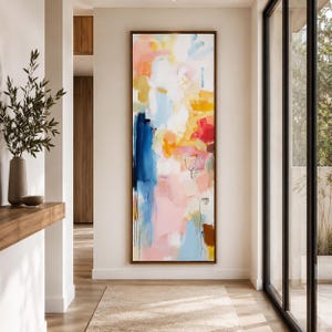 May include: An abstract painting in a dark wood frame. The artwork features a vibrant mix of colors, including blue, pink, yellow, and red, with brushstrokes and layered textures. The painting is displayed in a well-lit hallway.