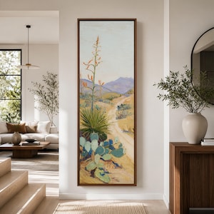 May include: A framed landscape painting depicting a desert scene with a winding dirt road, cacti, and a tall flowering plant. The artwork features a palette of muted greens, yellows, and blues, with a brown frame. The painting evokes a sense of the American Southwest.