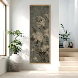 May include: A long, vertical framed canvas art piece featuring a floral design in shades of white, gray, and brown against a muted teal background. The artwork is displayed in a well-lit room with a large window and a potted plant.