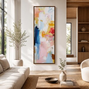May include: Abstract art painting in a vertical rectangular format. The artwork features a blend of colors, including blue, pink, yellow, and white, with a brown frame. The painting is displayed in a modern living room setting.