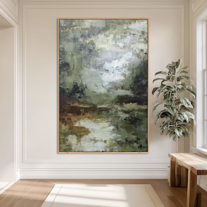 May include: Large abstract painting with a gold frame. The artwork features a landscape scene with shades of green, brown, and white. The brushstrokes create a textured effect, evoking a sense of depth and movement. The painting is displayed in a bright, neutral-toned room.