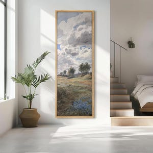 May include: A framed landscape painting with a gold-colored frame. The artwork depicts a field with trees, a path, and a cloudy sky. Blue flowers are visible in the foreground. The painting is displayed in a bright, modern room.
