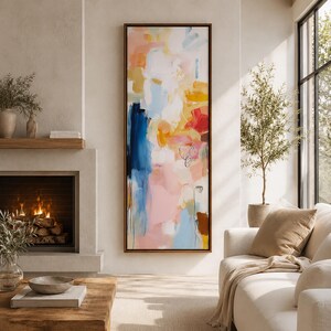May include: Abstract art painting in a dark wood frame. The artwork features a colorful composition of pink, blue, yellow, and red brushstrokes. The painting hangs above a fireplace in a living room setting.