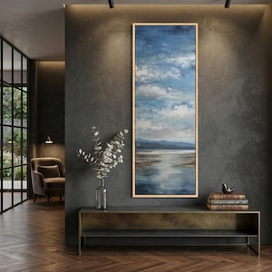 May include: Vertical landscape painting with a serene sky and water scene. The artwork features a blue sky with fluffy white clouds, reflecting in the calm water. The painting is framed in a light wood frame and hangs on a dark gray wall.