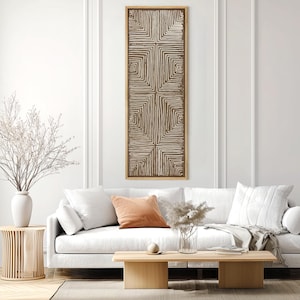 May include: A rectangular, vertical wall art piece with a geometric design. The artwork features a series of brown, square-like patterns on a cream-colored background, framed in a light brown wooden frame. The art hangs above a white sofa with decorative pillows.