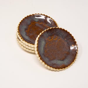 May include: A stack of six small, round ceramic plates with a scalloped edge. The plates have a dark brown center with a blue-green hue and a cream-colored rim. The plates are stacked on a white surface.