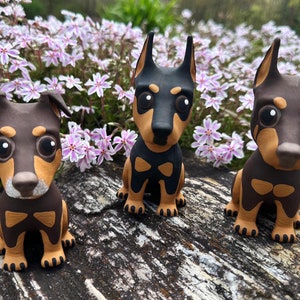 May include: Three small brown and tan dog figurines with black eyes and white noses. The figurines are sitting on a wooden surface.