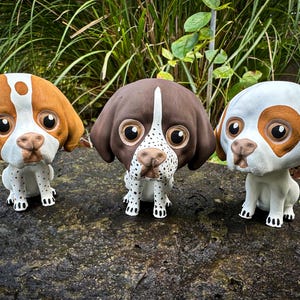 May include: Three ceramic figurines of dogs, each with a different color scheme. The dog on the left is brown and white, the middle dog is brown and white, and the dog on the right is white with brown spots. All three dogs have large, round eyes and are sitting with their paws tucked underneath them.