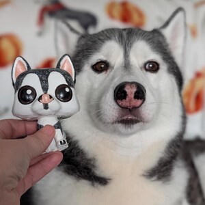 May include: A white and gray husky dog with black eyes looks at the camera. The dog is holding a small, white and gray figurine of a husky dog with large, black eyes.