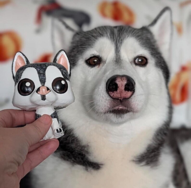 May include: A white and gray husky dog with black eyes looks at the camera. The dog is holding a small, white and gray figurine of a husky dog with large, black eyes.