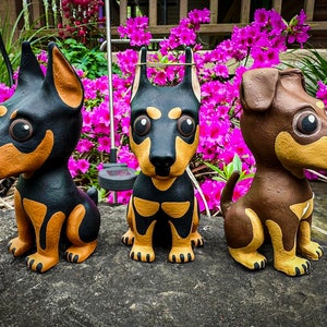 May include: Three small, hand-painted dog figurines. The dogs are black, brown, and tan with white accents. They are sitting on a gray, textured surface.